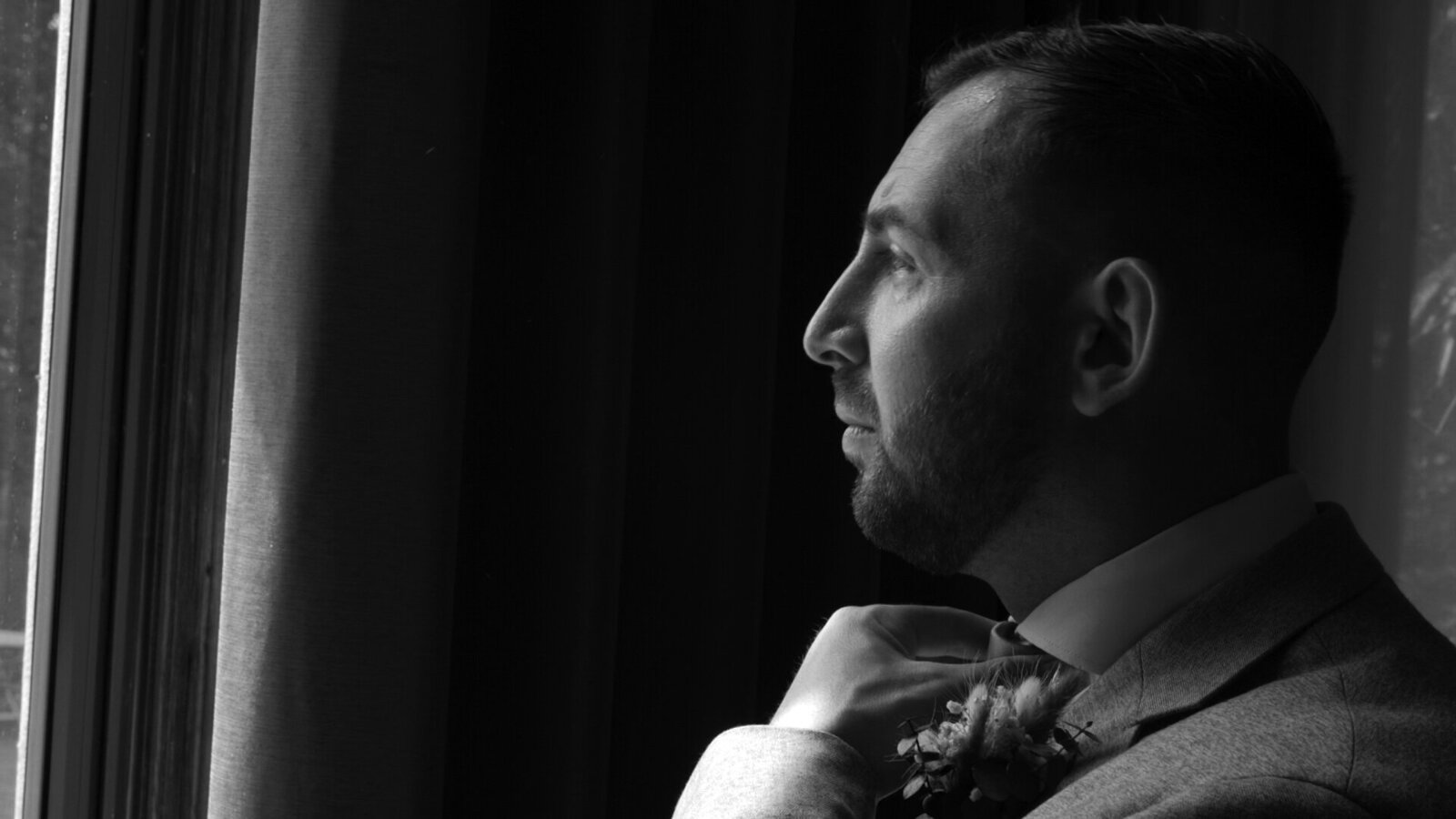 Black-and-white portrait of a groom adjusting his collar beside a window